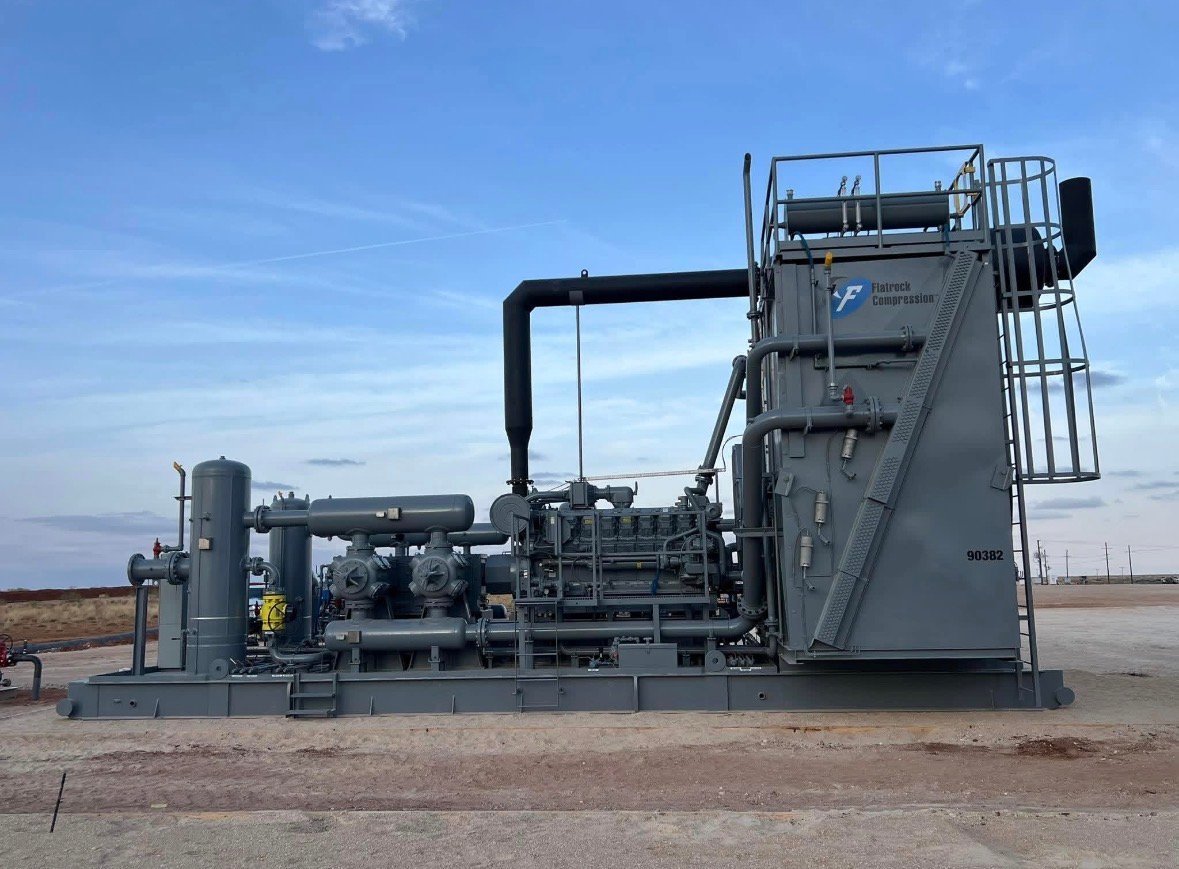 Large industrial compressor unit with gray metal components, piping, and control tower stationed on desert ground under clear blue sky